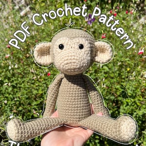 May include: A crocheted monkey toy in tan and cream colors. The monkey has black eyes and is sitting with its legs extended. The image includes the text "PDF Crochet Pattern" and "The Little Monkey". The background is a blurred view of green foliage.