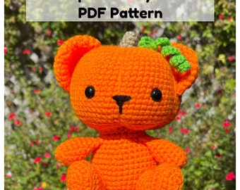 May include: A crocheted pumpkin teddy bear with a green stem and leaves. The bear is orange and has black eyes and a black nose.