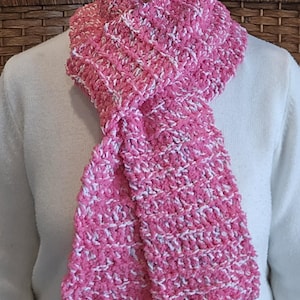 May include: A pink and white crocheted scarf is draped around a person's neck. The scarf is made of a soft, textured yarn, with a mix of pink and white threads. The scarf is a cozy accessory.