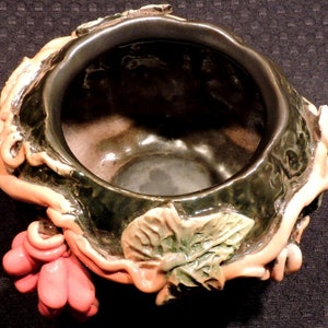 May include: A ceramic planter with a green glaze and a pink and orange vine design. The planter has a wide, round opening and a deep interior.