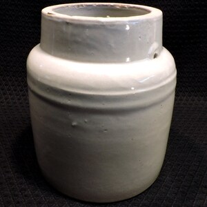 May include: White glazed ceramic crock with a round body and a wide opening. The crock has a smooth, slightly textured surface and a small hole on the side.