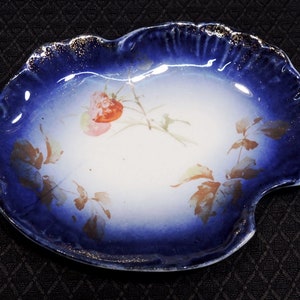 May include: A blue and white porcelain dish with a scalloped edge and a floral design. The dish is decorated with a gold rim and a floral pattern in shades of pink and brown.