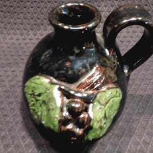 May include: A black ceramic pitcher with a handle and a spout. The pitcher features a green grape and leaf design on the front. The pitcher has a glossy finish and is likely used for serving beverages.