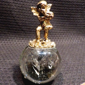 May include: A gold cherub figurine stands atop a clear glass orb with a textured surface. The cherub is holding a small object in its hands.