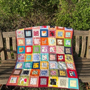 May include: A handmade patchwork quilt featuring numerous squares with diverse patterns and colours. The quilt, with a red border, is draped over a wooden bench. The squares include floral, animal, and abstract designs, creating a vibrant textile.