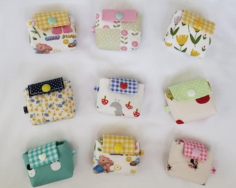 May include: Nine small, square fabric pouches with patterned designs. Each pouch has a different pattern, including floral, gingham, and cartoon characters. The pouches have a flap closure with a button.