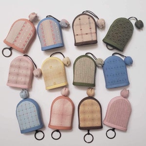 May include: A collection of eleven arched fabric key covers in various colors and patterns, including floral, plaid, and solid textures. Each cover features a small fabric ball charm attached by a cord and a metal ring at the base for keys.