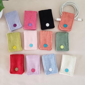May include: A collection of corduroy cable organizers in various colors, including pink, black, and green. Each organizer has a button in a contrasting color. A white cable is placed on the top right.