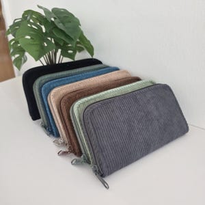 May include: A collection of six rectangular zip-around wallets made of corduroy fabric, arranged in a row. The wallets are available in black, teal, blue, beige, brown, light green, and dark gray, each featuring a metal zipper closure.