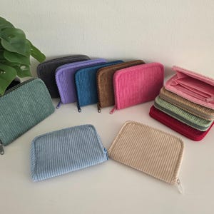 May include: A collection of corduroy wallets in various colors, including green, blue, purple, brown, and pink. Some wallets are closed, while others are open, revealing interior compartments. The wallets are arranged on a white surface.