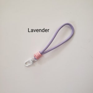 K&ouml;nnte beinhalten: Ein Lavendel-Telefonband mit einem rosa Knoten und einem wei&szlig;en Metallclip. Das Band tr&auml;gt den Schriftzug "Lavender" in schwarzer Schrift.