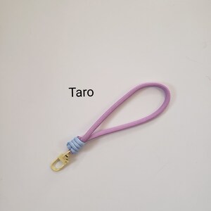 K&ouml;nnte beinhalten: Ein hellviolettes und blaues Lanyard mit einem goldenen Metallverschluss. Das Lanyard tr&auml;gt den Schriftzug "Taro".