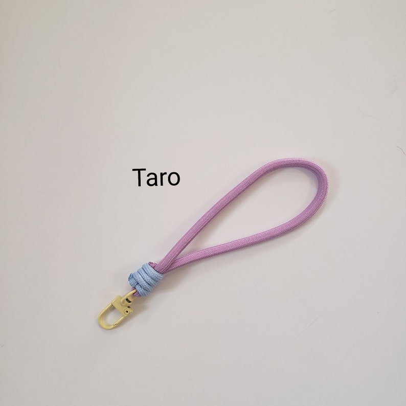 K&ouml;nnte beinhalten: Ein hellviolettes und blaues Lanyard mit einem goldenen Metallverschluss. Das Lanyard tr&auml;gt den Schriftzug "Taro".