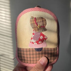May include: A small, oval-shaped fabric pouch featuring a quilted applique of a figure wearing a plaid bonnet and a red floral dress. The pouch is bordered by a red and white gingham trim, with a brown plaid fabric panel at the bottom base.