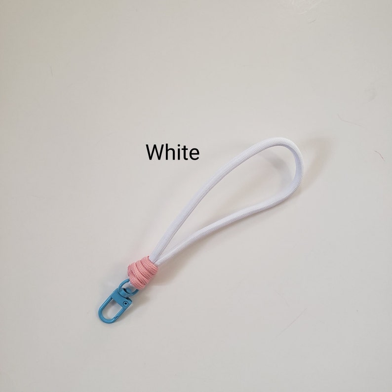 K&ouml;nnte beinhalten: Wei&szlig;es Lanyard mit einem rosa-blauen Knoten und einem blauen Metallclip. Das Lanyard tr&auml;gt die Aufschrift "White" auf dem wei&szlig;en Teil.