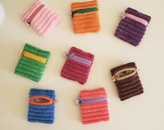 May include: A collection of small, corduroy zippered pouches in various colors, including pink, orange, green, burgundy, blue, and red. Each pouch has a contrasting zipper and a floral fabric accent. The pouches are arranged on a light-colored surface.