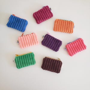 May include: A collection of small, rectangular corduroy zipper pouches in various colors, including blue, pink, purple, orange, green, and brown. Each pouch has a matching zipper and a small tag attached. The pouches are arranged on a white surface.