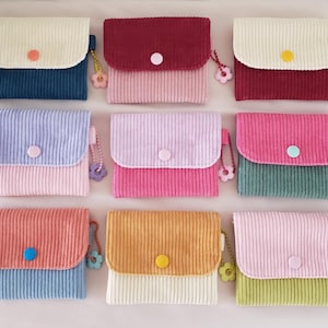May include: Ten small, colorful wallets with a corduroy texture. Each wallet has a different color combination and a small button closure. The wallets are arranged in two rows of five.