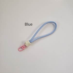 K&ouml;nnte beinhalten: Ein hellblaues Lanyard mit einem wei&szlig;en und cremefarbenen Knoten und einem rosafarbenen Metallclip. Das Lanyard tr&auml;gt den Schriftzug "Blue" in Schwarz.