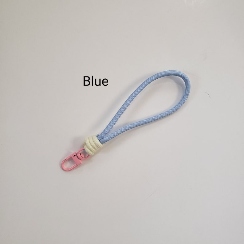 K&ouml;nnte beinhalten: Ein hellblaues Lanyard mit einem wei&szlig;en und cremefarbenen Knoten und einem rosafarbenen Metallclip. Das Lanyard tr&auml;gt den Schriftzug "Blue" in Schwarz.