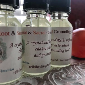 May include: Three clear glass bottles with dropper tops and white labels. The labels have text that reads "Root & Sacroot & Sacral Grounding Oil". The text on the labels describes the oil as a crystal and Reiki infused chakra activation and grounding tool.