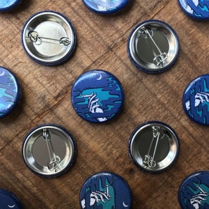 May include: Several round, blue pin badges with a night-themed illustration. The design shows a hand holding a cigarette against a dark blue backdrop with a crescent moon and stars. The badges are arranged on a wooden surface.