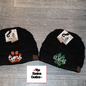 May include: Two black knit beanies with a paw print and team name embroidered on each. The beanie on the left has an orange glitter paw print and says "Tigers". The beanie on the right has a green glitter paw print and says "Wolves".