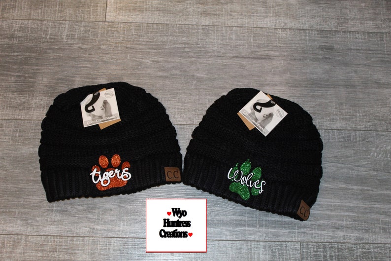 May include: Two black knit beanies with a paw print and team name embroidered on each. The beanie on the left has an orange glitter paw print and says "Tigers". The beanie on the right has a green glitter paw print and says "Wolves".