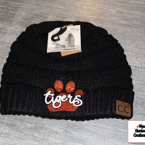 May include: Black knit beanie with a brown glitter paw print and the word "tigers" in white script. The beanie has a brown leather patch with the letters "CC" on it.