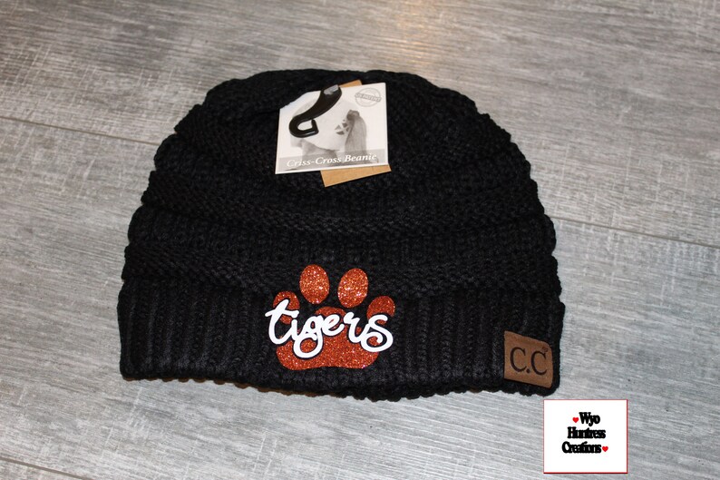 May include: Black knit beanie with a brown glitter paw print and the word "tigers" in white script. The beanie has a brown leather patch with the letters "CC" on it.