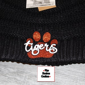 May include: Black knitted beanie with a brown glitter paw print and the word "tigers" in white lettering. The beanie has a brown leather patch with the letters "C.C."