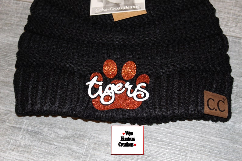May include: Black knitted beanie with a brown glitter paw print and the word "tigers" in white lettering. The beanie has a brown leather patch with the letters "C.C."