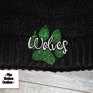 May include: Black knit beanie with a green glitter paw print and the word "Wolves" in white cursive lettering.