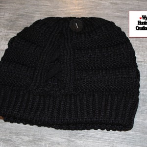 May include: Black knitted beanie with a cable knit pattern. The beanie has a fold-over cuff and a small tag with the text "Wyo Huntress Creations".