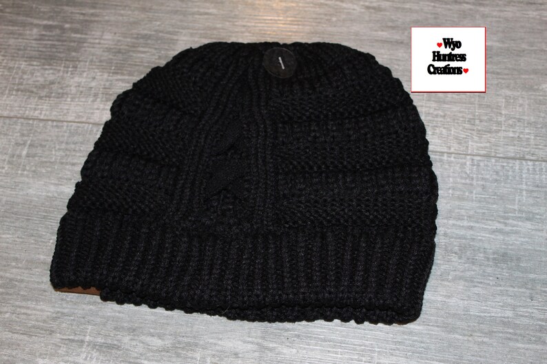May include: Black knitted beanie with a cable knit pattern. The beanie has a fold-over cuff and a small tag with the text "Wyo Huntress Creations".