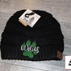 May include: Black knit beanie with a green glitter paw print and the word "Wolves" in white. The beanie has a brown leather patch with the letters "CC" on it.