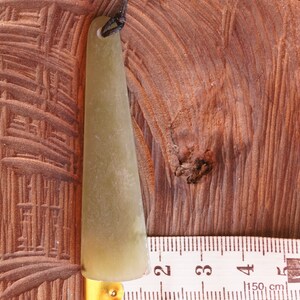 Puede incluir: Una herramienta de gua sha de jade verde claro con un agujero en la parte superior para una cuerda. La herramienta est&aacute; sobre una superficie de madera junto a una regla que dice "Birch 150 cm".