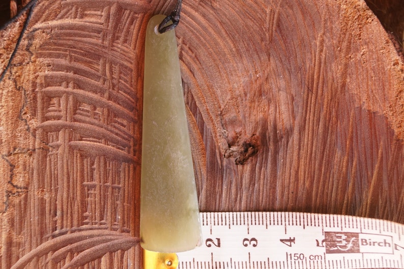 Puede incluir: Una herramienta de gua sha de jade verde claro con un agujero en la parte superior para una cuerda. La herramienta est&aacute; sobre una superficie de madera junto a una regla que dice "Birch 150 cm".