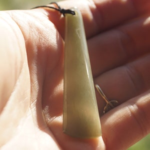 Puede incluir: Un colgante de jade verde con una superficie lisa y pulida cuelga de un cord&oacute;n negro. El colgante tiene la forma de un rect&aacute;ngulo largo y delgado con bordes redondeados.