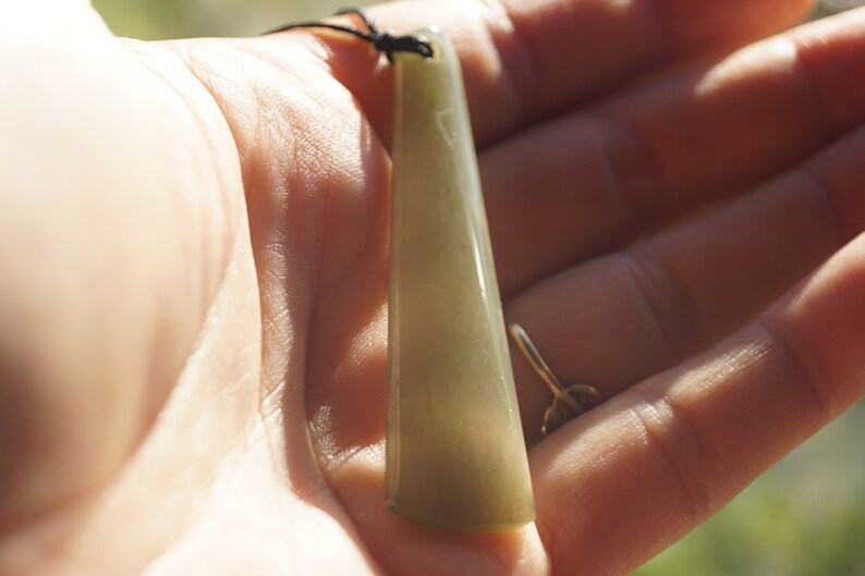 Puede incluir: Un colgante de jade verde con una superficie lisa y pulida cuelga de un cord&oacute;n negro. El colgante tiene la forma de un rect&aacute;ngulo largo y delgado con bordes redondeados.
