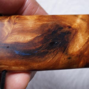 May include: A close-up of a wooden pendant with a dark brown and light brown wood grain pattern. The pendant has a blue streak running through it and is attached to a black cord.