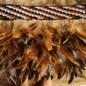 Könnte beinhalten: Nahaufnahme eines traditionellen Kleidungsstücks mit einem gewebten Band mit einem schwarzen, weißen und braunen Zickzackmuster. Darunter befindet sich eine dichte Anordnung brauner und hellbrauner Federn.