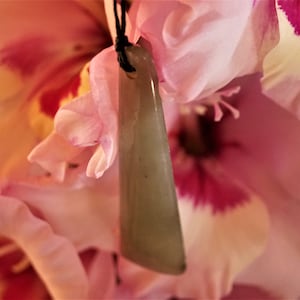 Puede incluir: Un colgante de jade verde claro con una superficie lisa y pulida cuelga de un cord&oacute;n negro. El colgante tiene forma de tri&aacute;ngulo largo y delgado. El colgante est&aacute; apoyado sobre una flor rosa.