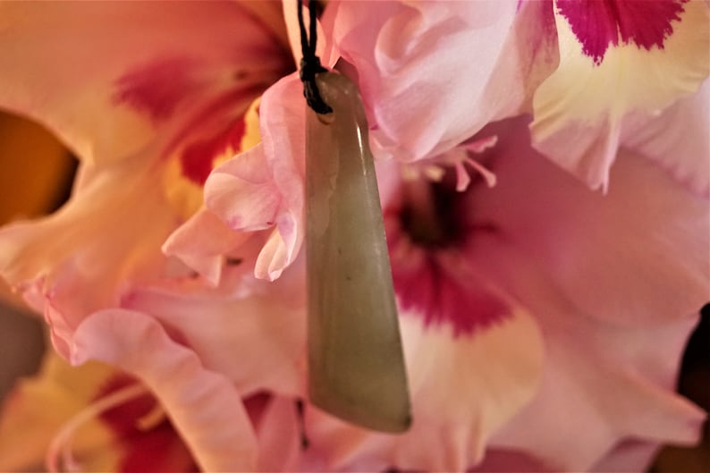 Puede incluir: Un colgante de jade verde claro con una superficie lisa y pulida cuelga de un cord&oacute;n negro. El colgante tiene forma de tri&aacute;ngulo largo y delgado. El colgante est&aacute; apoyado sobre una flor rosa.