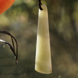 Puede incluir: Un colgante de jade verde claro con una superficie lisa y pulida. El colgante est&aacute; unido a un cord&oacute;n negro.