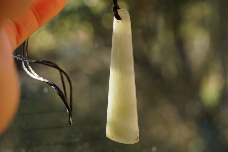 Puede incluir: Un colgante de jade verde claro con una superficie lisa y pulida. El colgante est&aacute; unido a un cord&oacute;n negro.