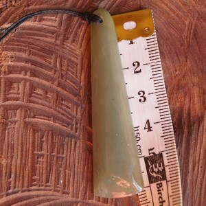 Puede incluir: Un colgante de jade verde claro con una punta redondeada, colgando de un cord&oacute;n negro. El colgante mide aproximadamente 12,5 cm de largo y est&aacute; al lado de una regla.