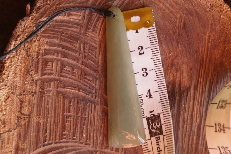 Puede incluir: Un colgante de jade verde claro con una punta redondeada, colgando de un cord&oacute;n negro. El colgante mide aproximadamente 12,5 cm de largo y est&aacute; al lado de una regla.