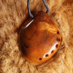 May include: A handcrafted wooden pendant with a natural, polished finish. The pendant is a warm brown color with a smooth surface and several small, dark holes. It hangs from a black cord, showcasing a unique, organic design.