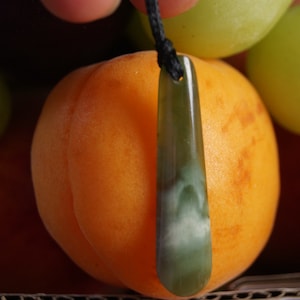 May include: A green jade pendant with a white swirl pattern hangs from a black cord. The pendant is resting on a peach colored fruit.